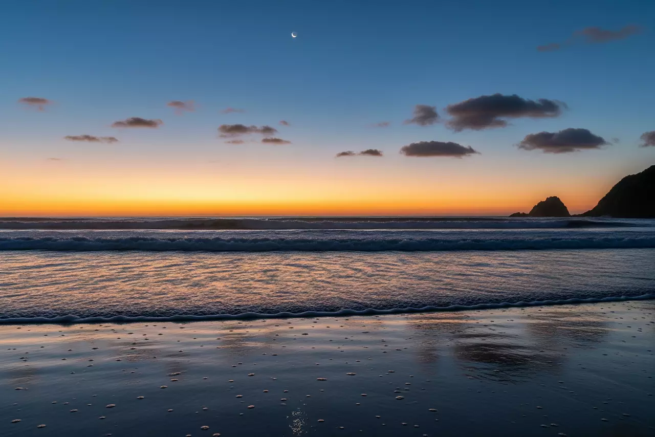 La tranquilidad del océano al atardecer, ideal para relajar la mente antes de dormir. Playa al anochecer con olas suaves rompiendo en la orilla y una luna tenue en el cielo.