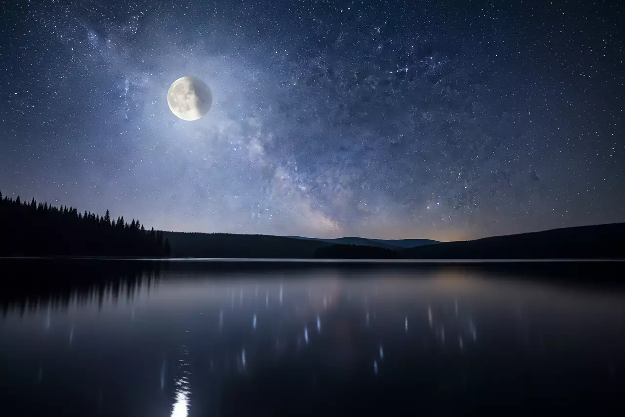 Un lago tranquilo bajo un cielo nocturno lleno de estrellas y la Vía Láctea, con la luna llena reflejándose en el agua y un bosque oscuro al fondo.